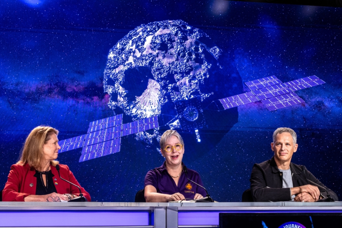 A woman speaks at a table with an image of an asteroid and spacecraft behind her
