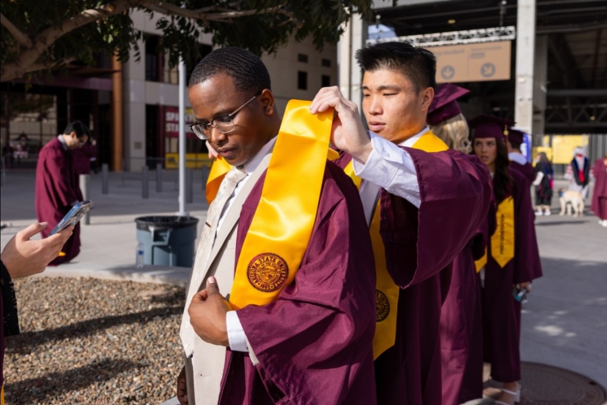 Graduate helping other graduate adjust gold sash before commencement