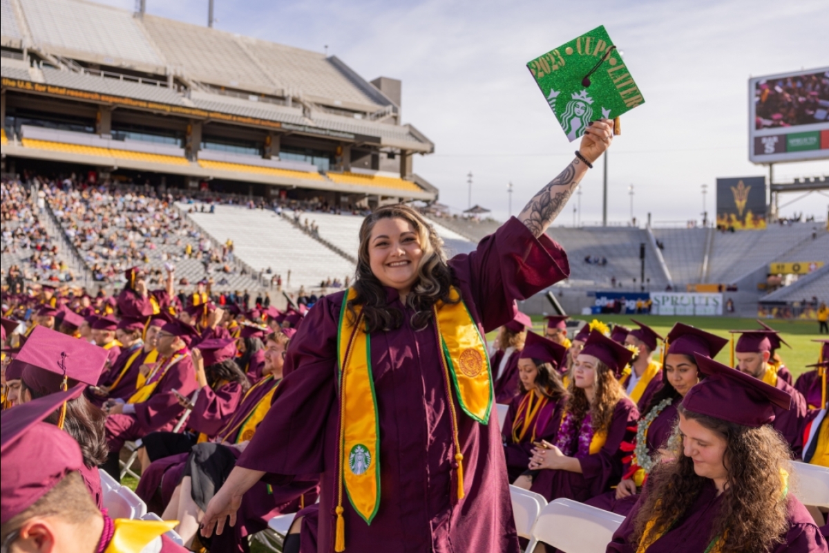 ASU grad in maroon cap and gown holds a green decorated graduation cap up in air