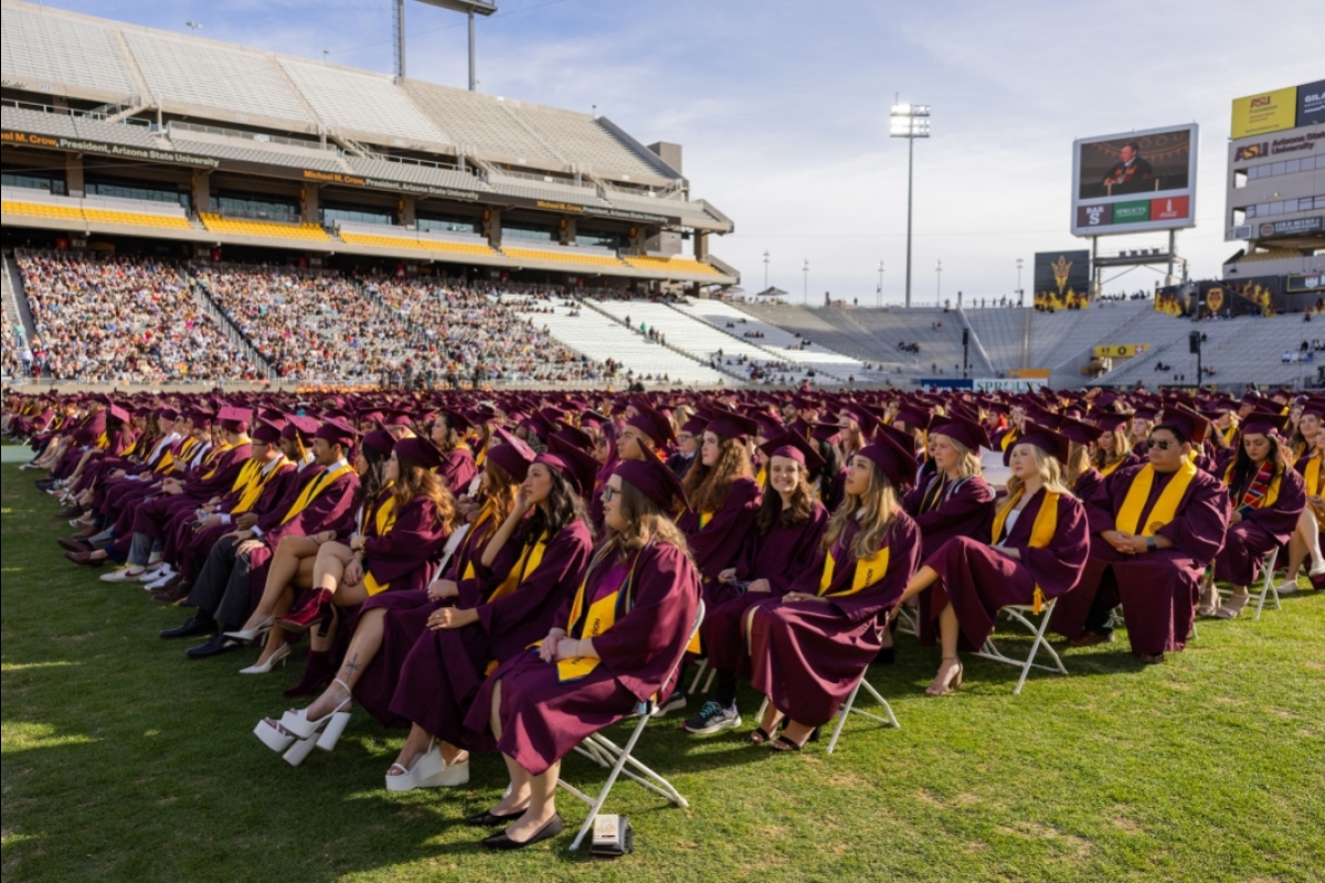 Rows of graduates sit on the football during Undergraduate Commencement with their families in the stands