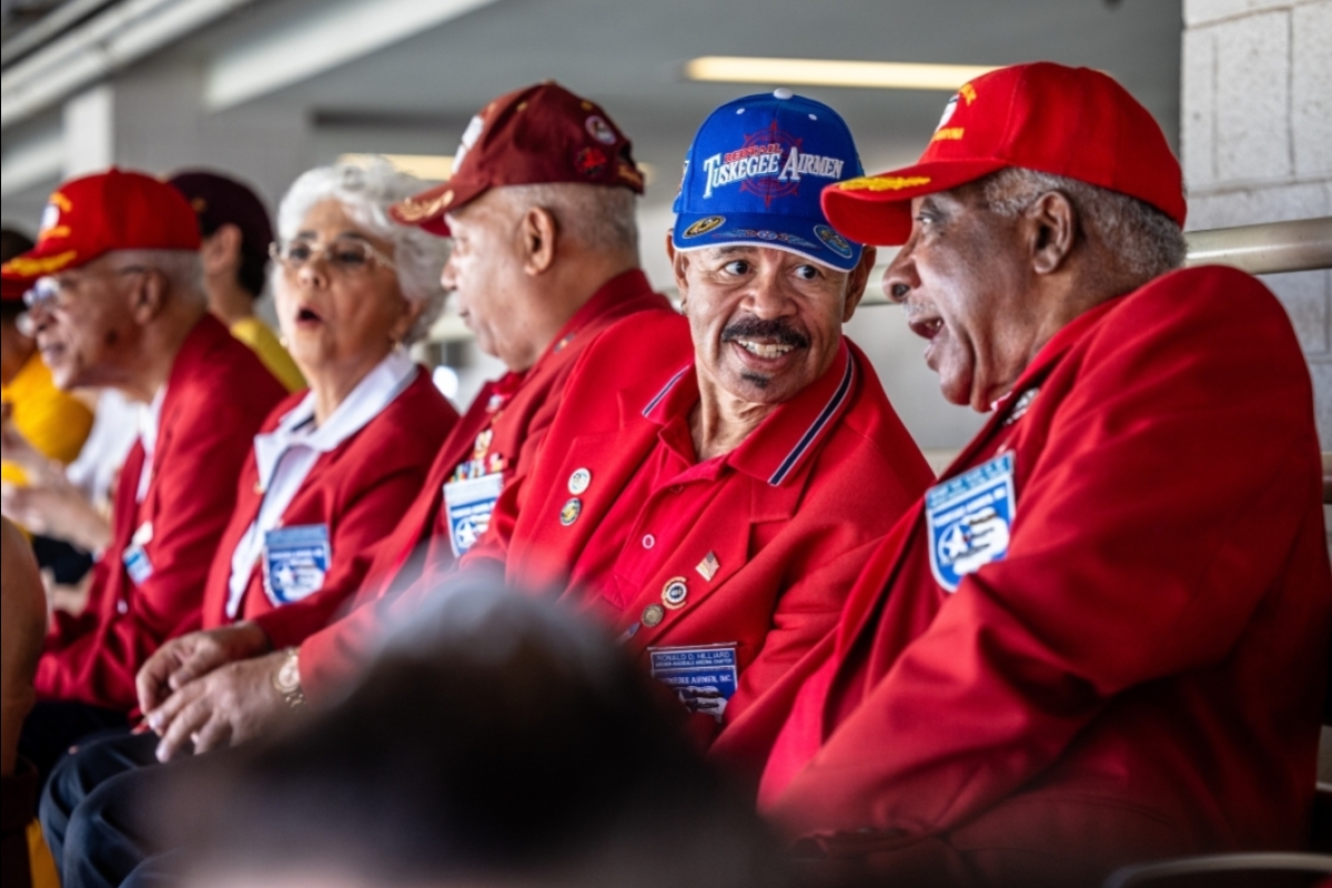 Older men in Tuskegee Airmen hats chat while sitting in stadium seats.