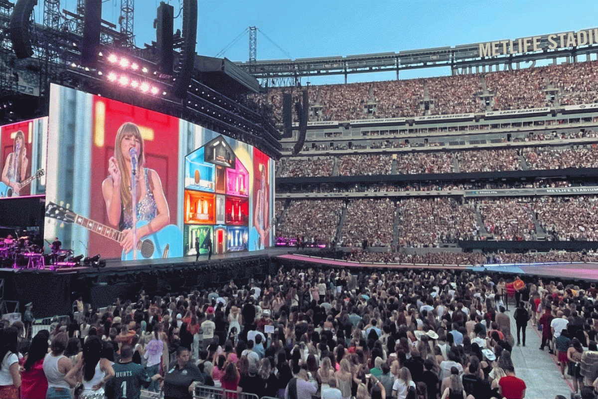 Fans fill a large arena in which a jumbotron displays the image of American singer-songwriter Taylor Swift.