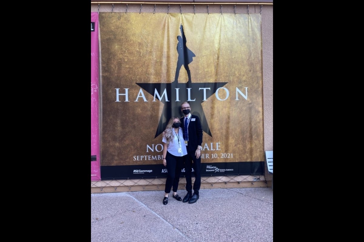 Portrait of two people posing in front of Hamilton poster