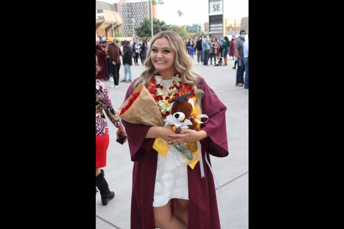 Woman posing at graduation in maroon gown holding flowers and a stuffed animal