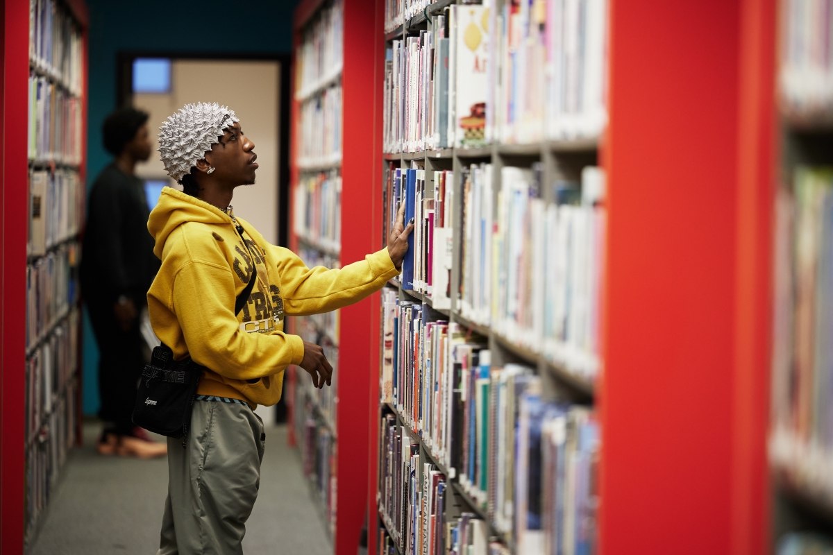 A student stands in front of a shelf of books in a library aisle.