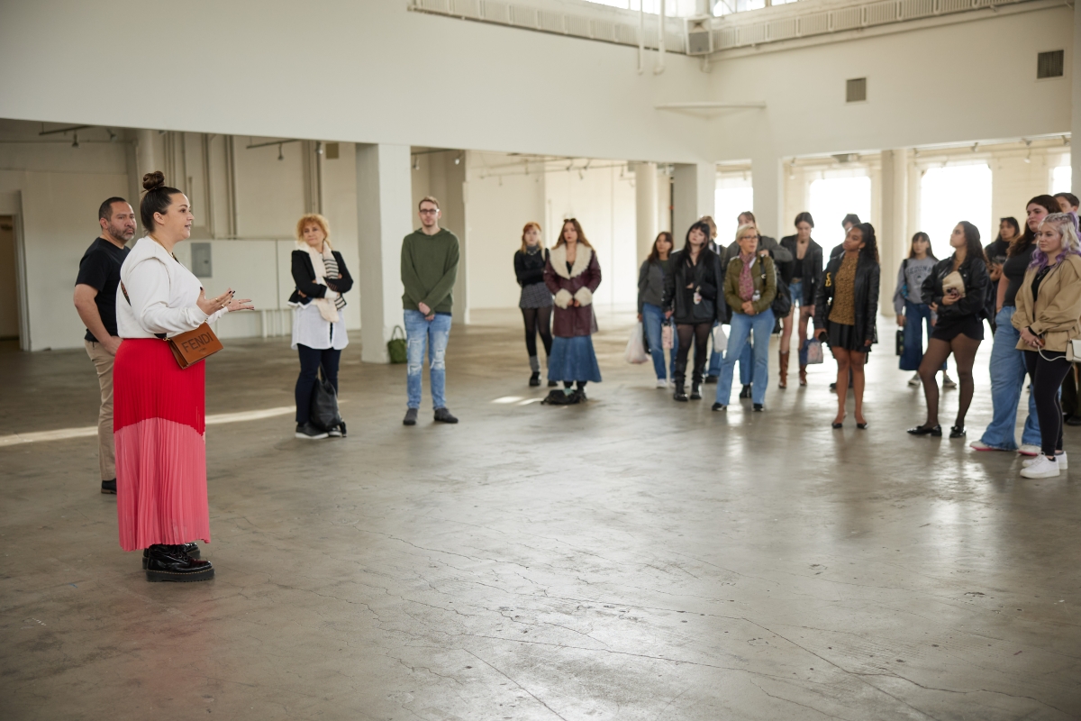 A woman stands in front of a group talking in a big, open interior space with concrete floors.