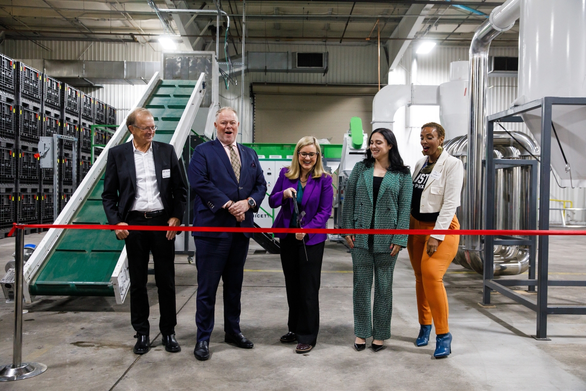 Schlosser, O'Neal, Gallego, Ansari and Waddell stand in front of a red ribbon with microfactory equipment behind them while Gallego cuts the ribbon