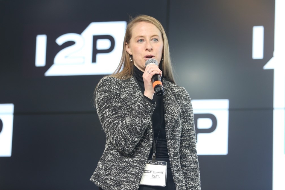 A woman speaking to an unseen audience in front of a video wall with I2P logos