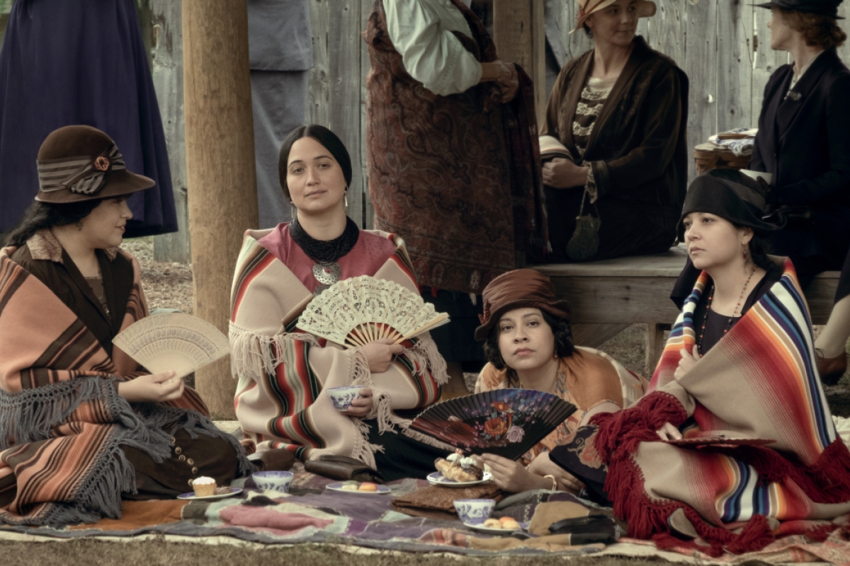 Women in old-fashioned and Indigenous-inspired clothing sitting on the ground holding folding fans