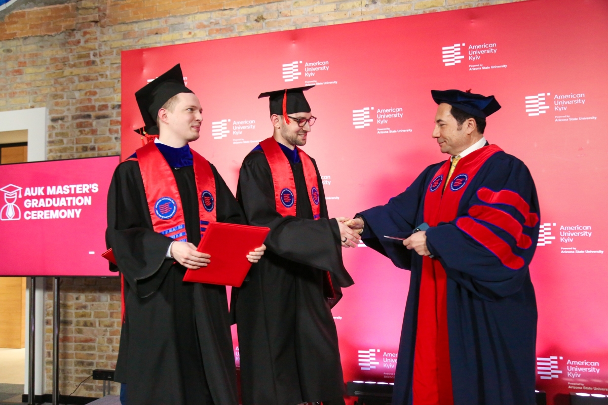 A student wearing a black graduation gown and red stole accepts a diploma from a similarly-dressed man.