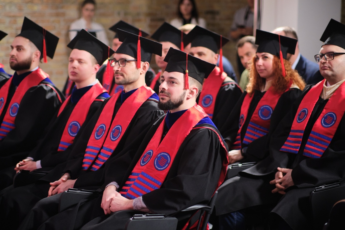 People wearing black graduation robes and red stoles seated in an audience.