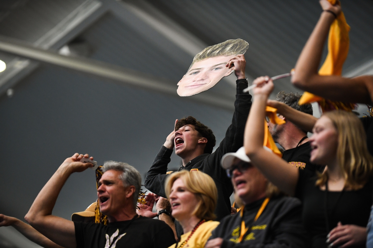 People in maroon and gold cheer in the stands holding a giant face cutout of a swimmer
