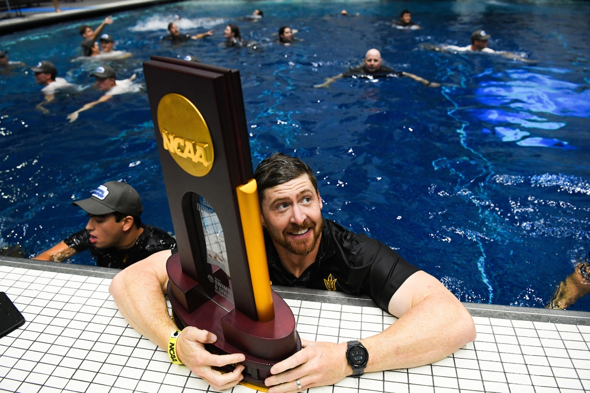 A coach in a pool hugs the NCAA trophy sitting on the pool deck.