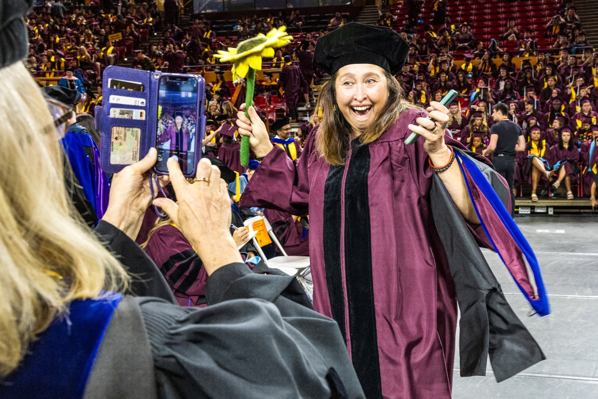 Woman in maroon graduation robe holding up fake sunflower while posing for a cellphone photo
