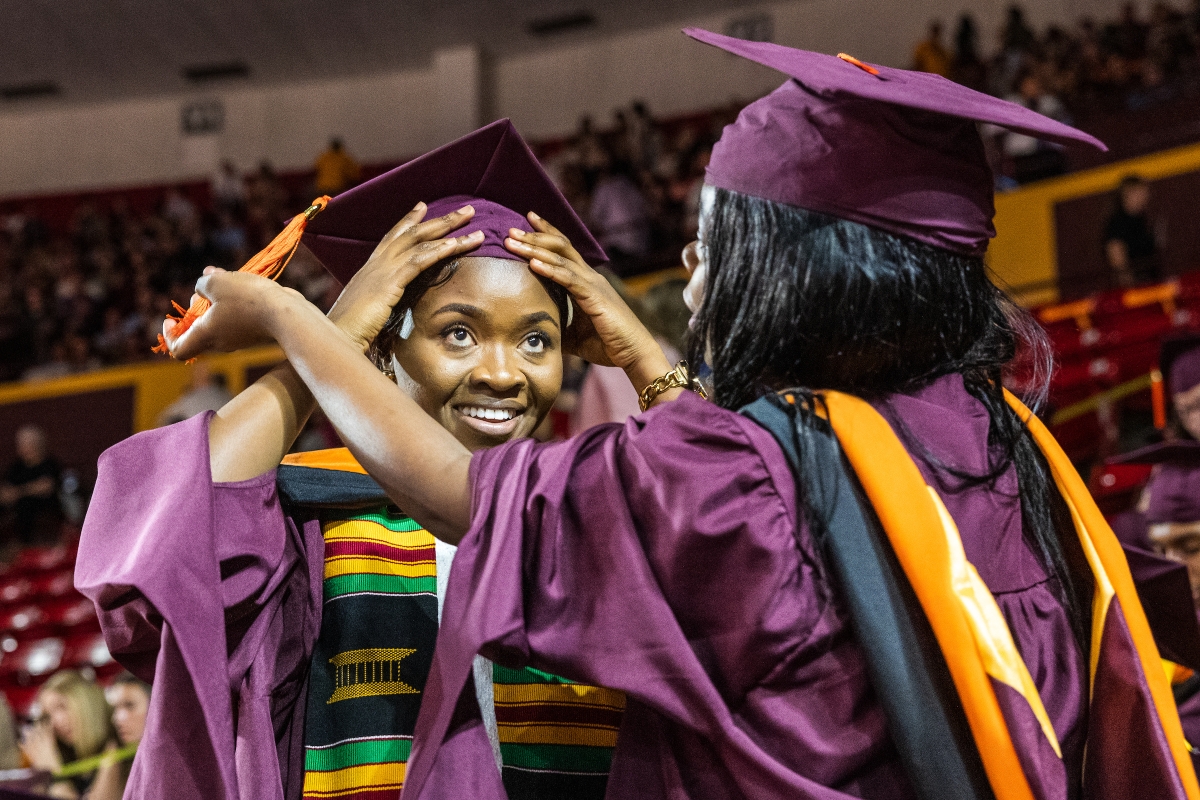 ASU graduate getting help adjusting her maroon cap from fellow graduate during commencement