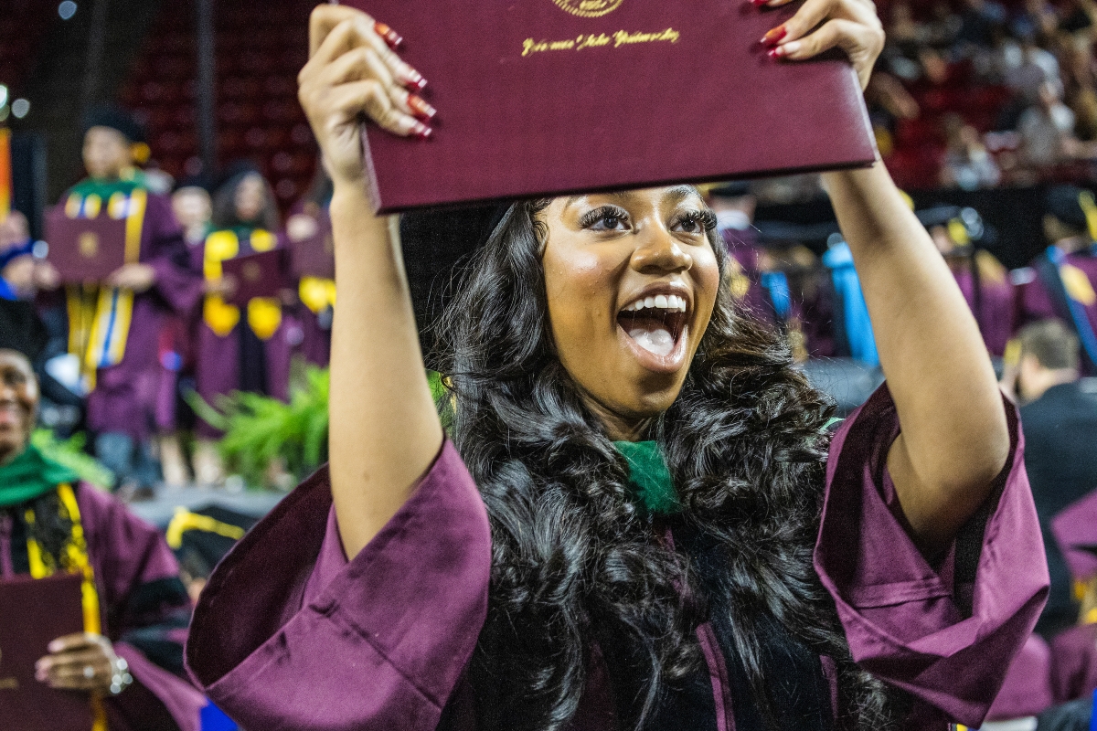 Black woman in maroon graduation gown with long dark hair holds up diploma during commencement