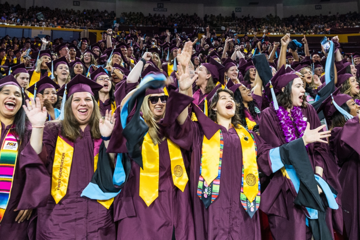Large group of graduates in maroon gowns raise arms in celebration during commencement