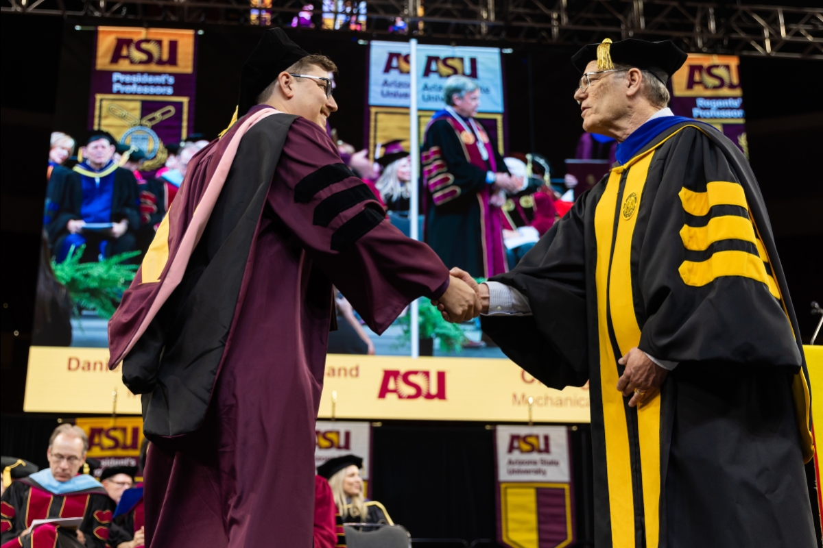 Student in maroon graduation robe shakes hands with dean in black and gold graduation robe
