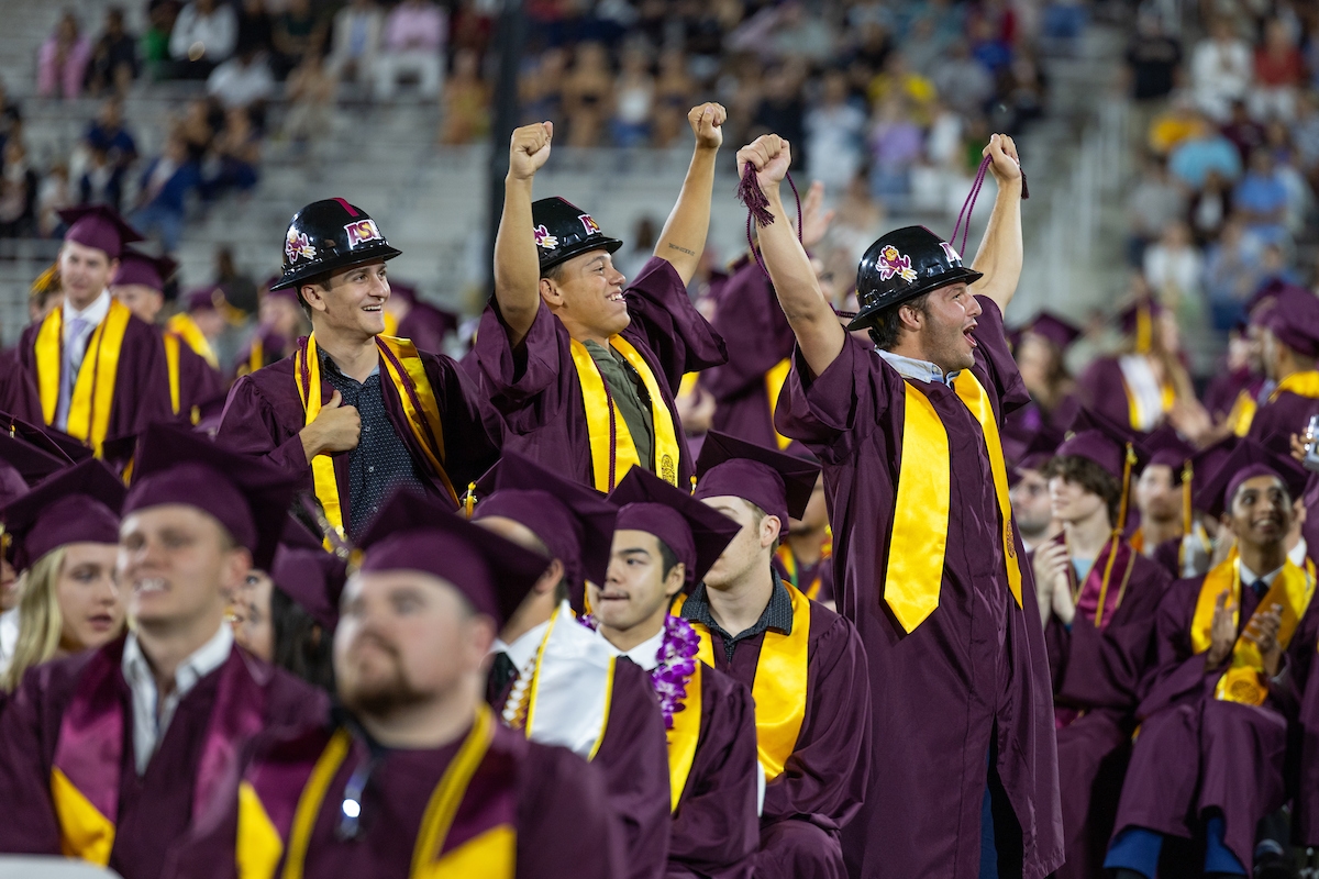 Three ASU graduates wearing maroon robes and construction hats stand and raise arms in the air during graduation