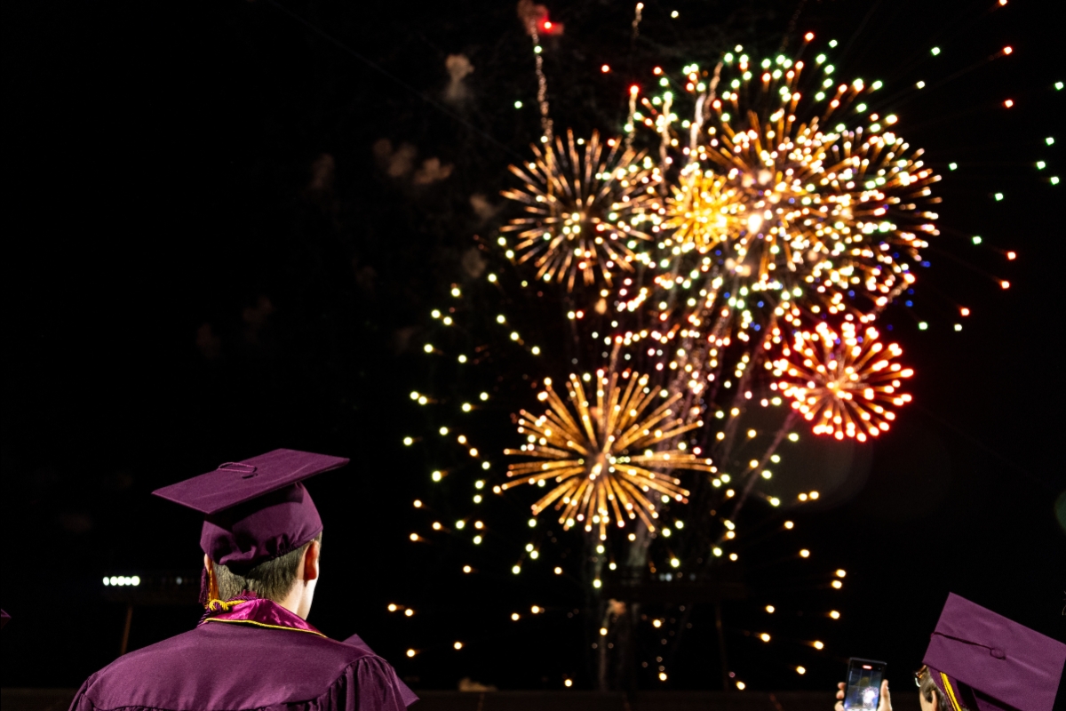 Graduates in maroon caps and gowns watch fireworks in the sky