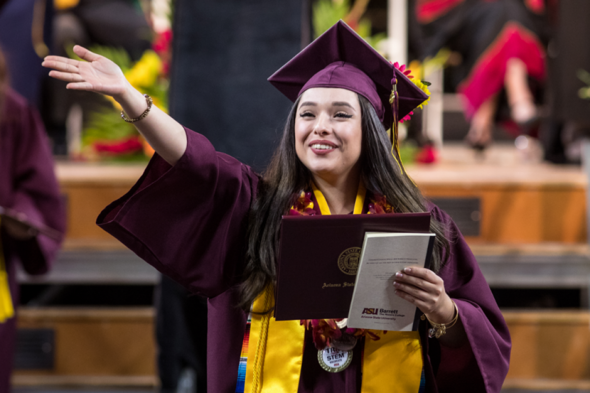 An ASU graduate holding a diploma cover waves at the audience as she walks at graduation