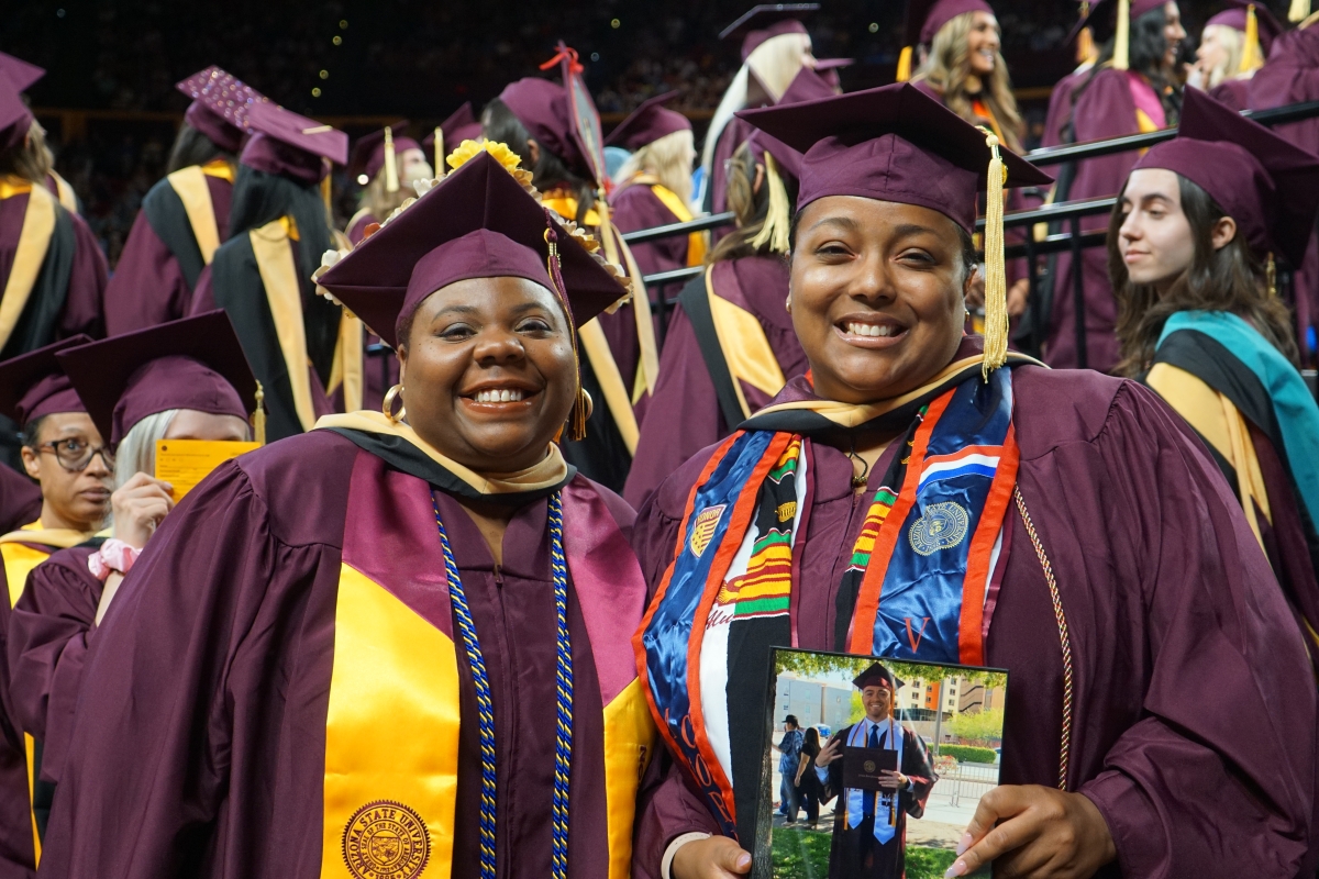 Two smiling graduates display a photo of a third graduate