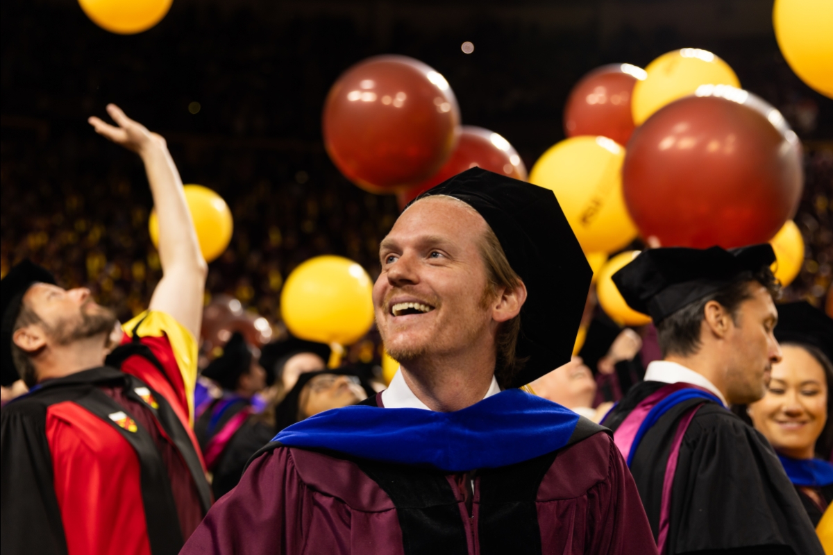 Man in graduation gown smiles with other graduates surrounding him and balloons falling in the background
