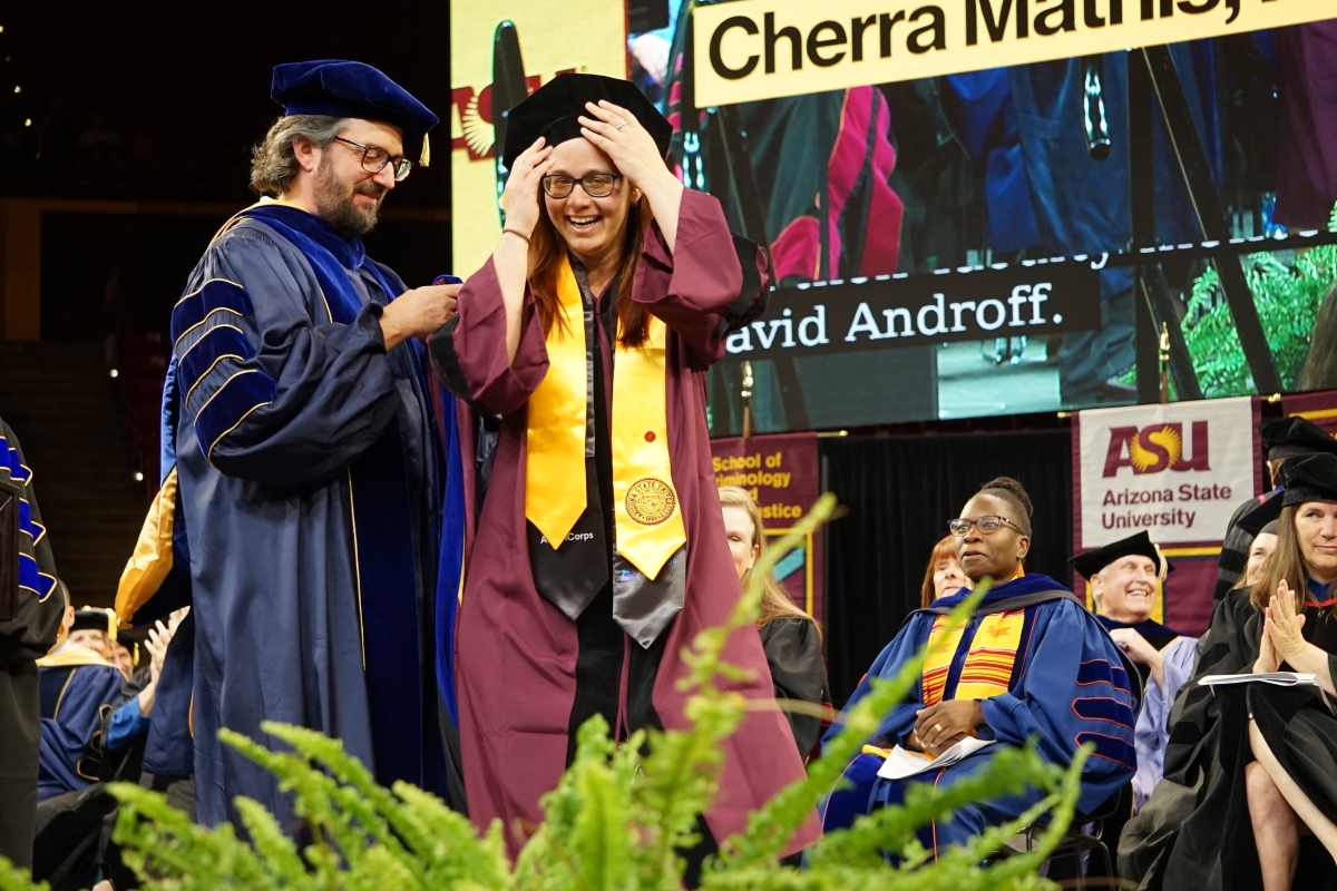 Woman smiles and holds onto her graduation cap on stage surrounded by others.