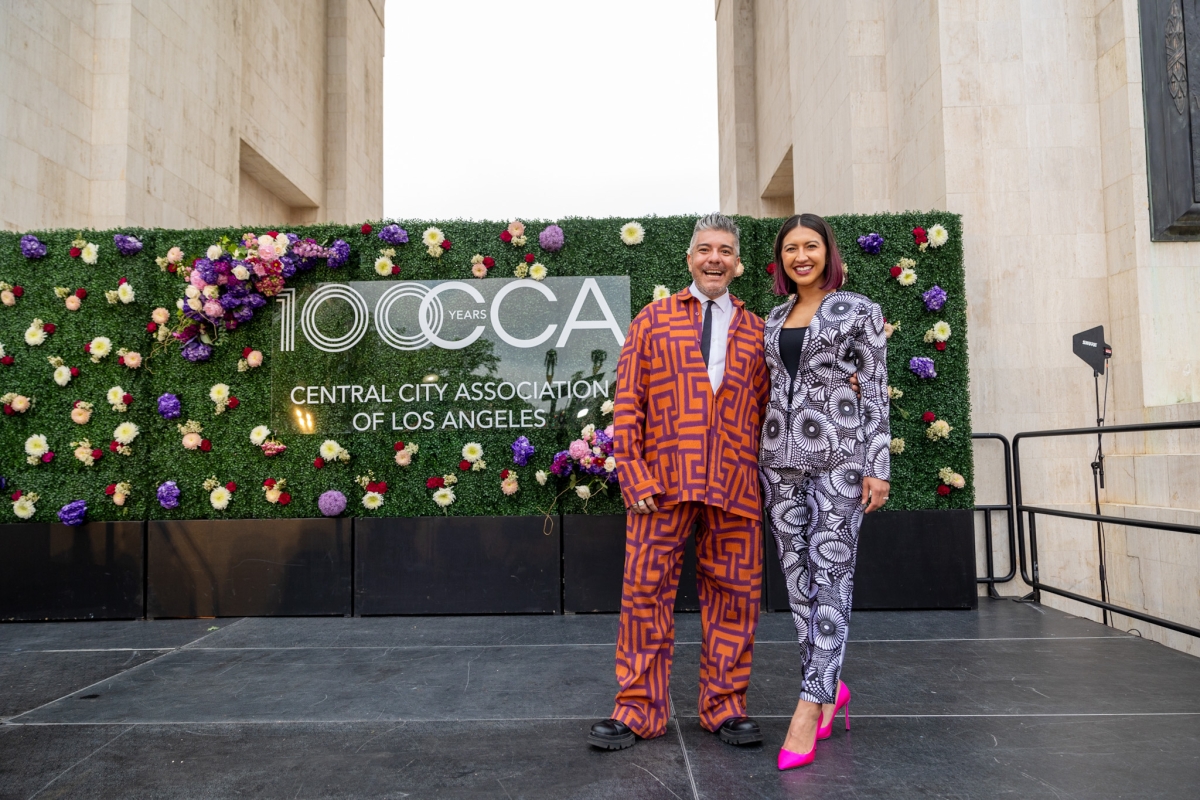 Man and woman wearing brightly colored and patterned clothes pose on a stage in front of a floral background.