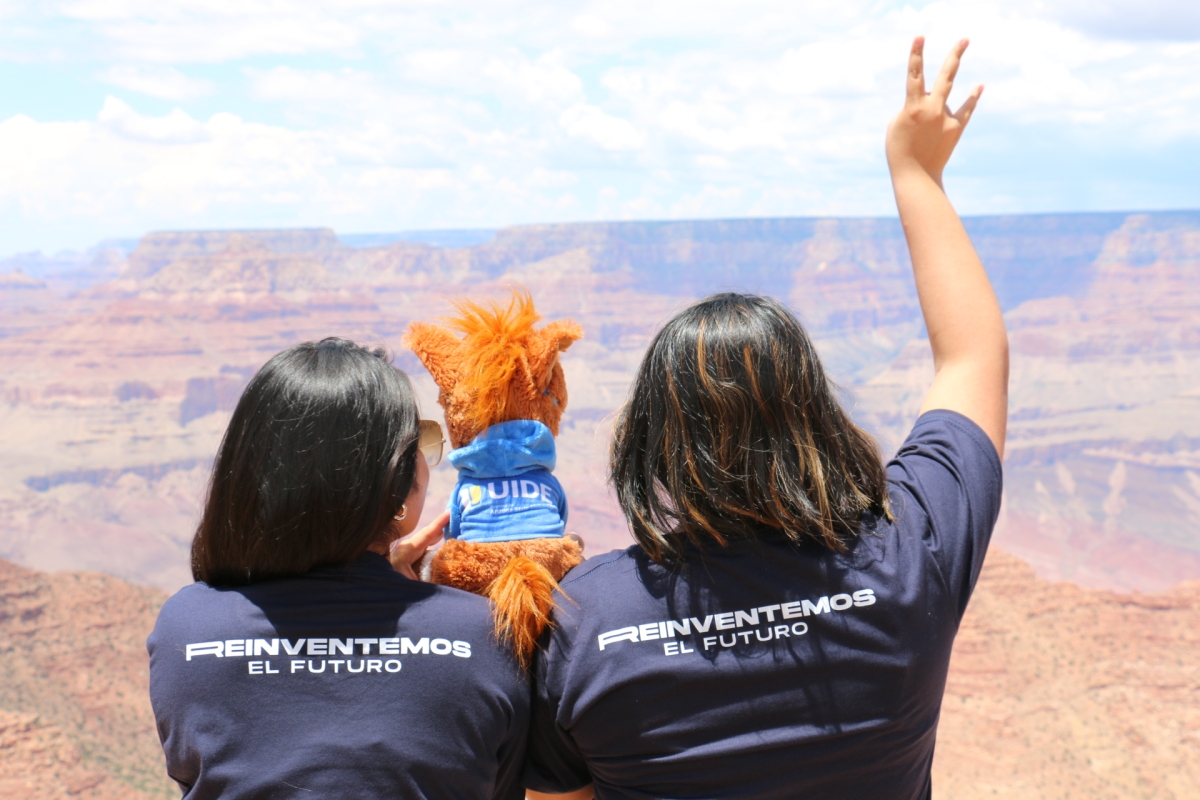 Two women seen from behind making an ASU pitchfork symbol with their hands as they look out at the Grand Canyon.