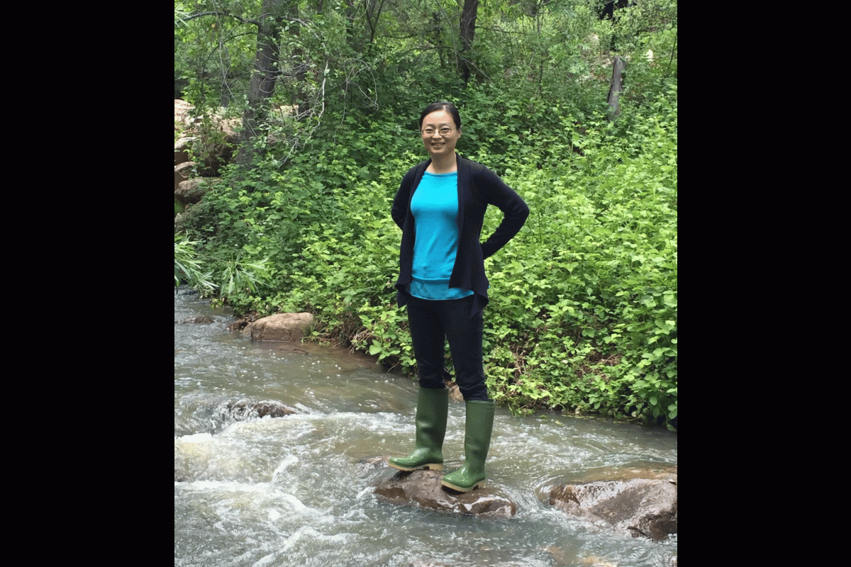 A woman in rubber boots stands in the middle of a stream in the woods