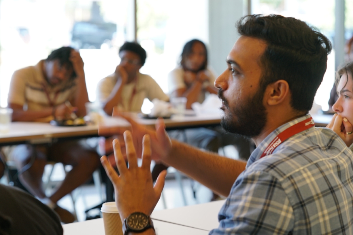 A group of students sit around desks in a circle for a discussion with the focus on a male student wearing a blue plaid shirt