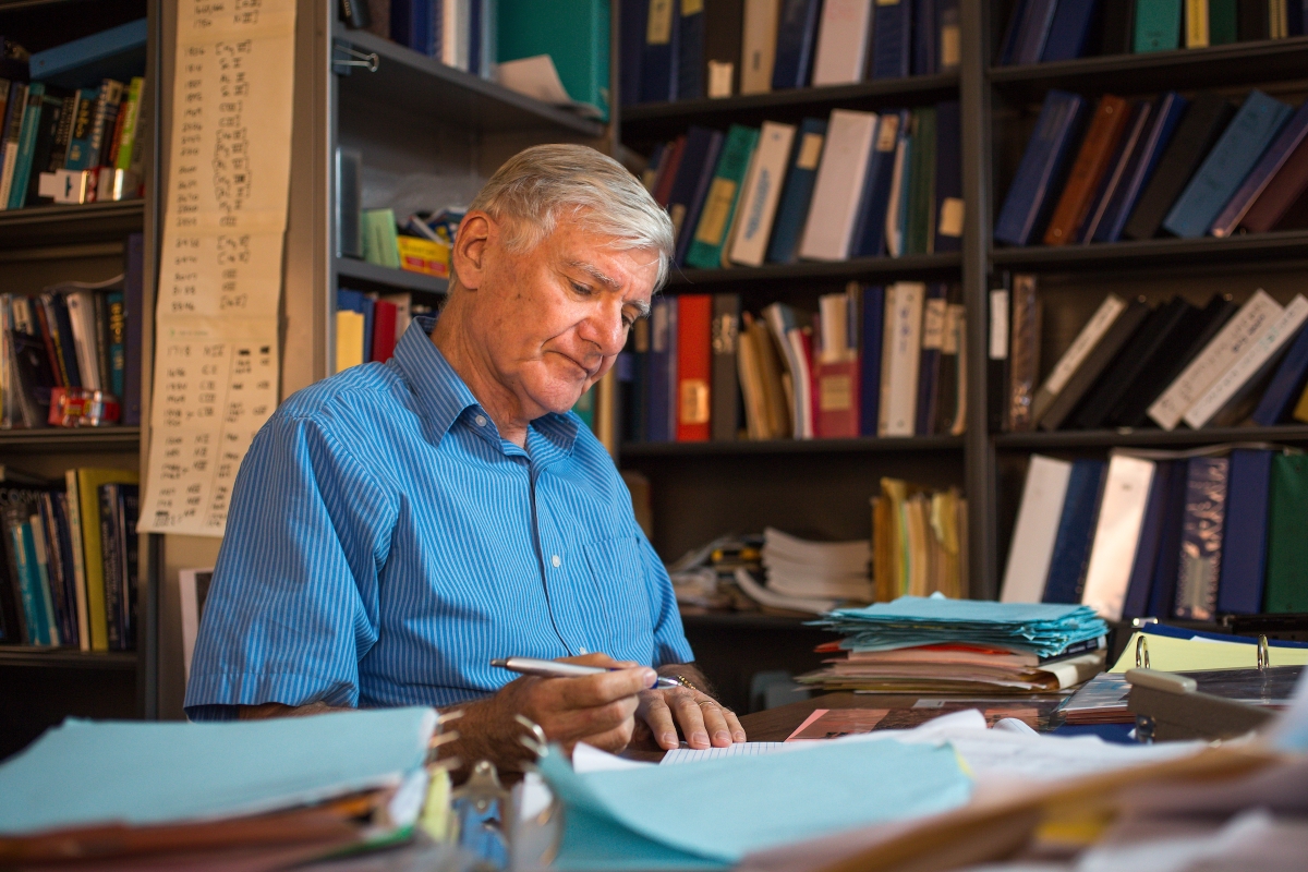 Older man wearing a blue button down shirt sits at a desk in his office with bookshelves behind him