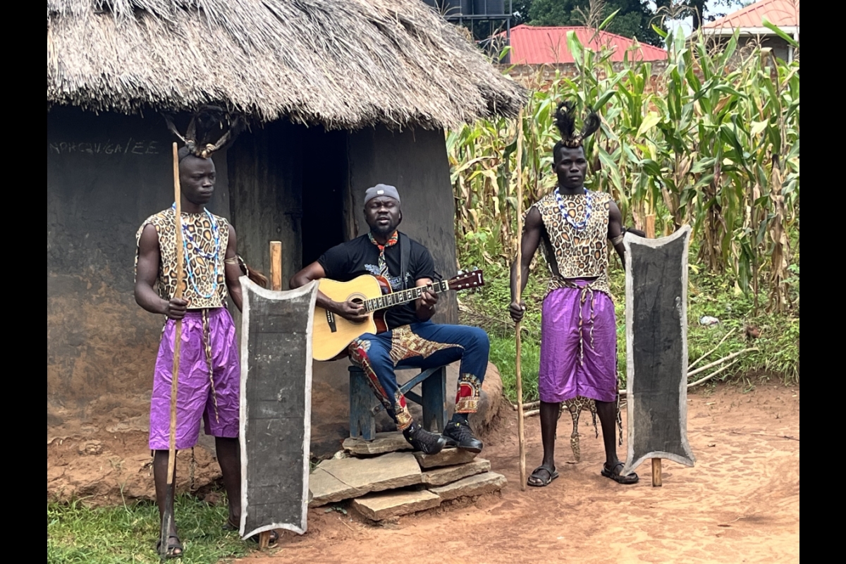 A man holding a guitar sings while two African warriors stand on either side of him