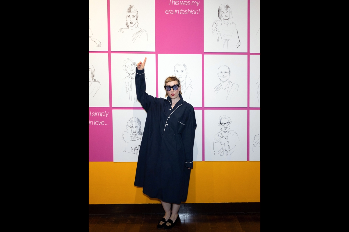 Woman in a black button-up ensemble poses in front of a wall with sketched portraits on it.