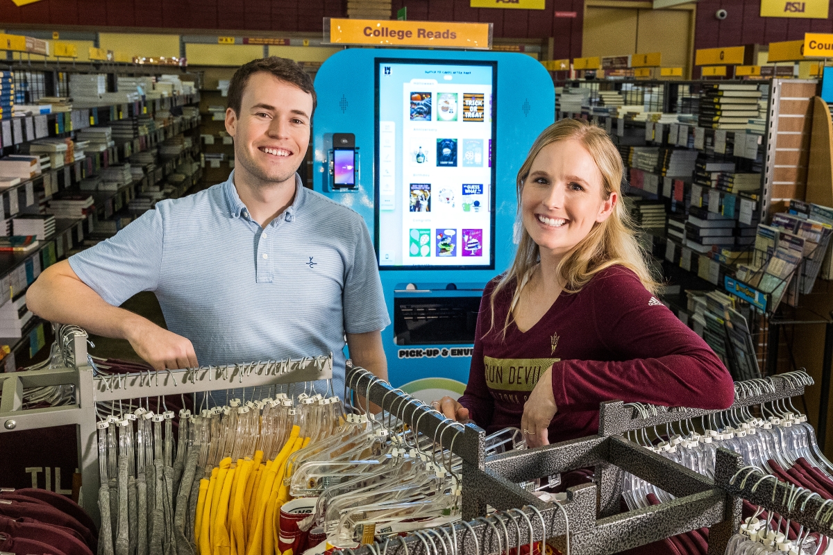 A man and woman smile in front of a kiosk