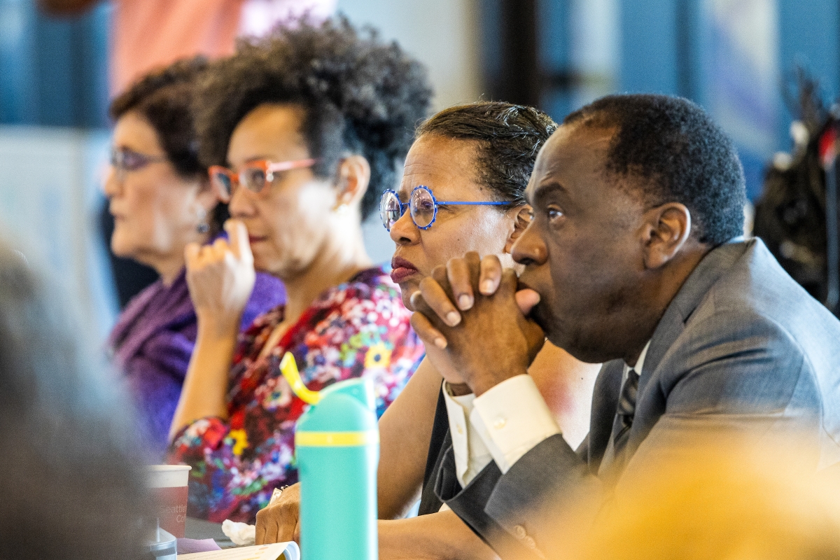 Four people of color sitting at a table listening to speakers