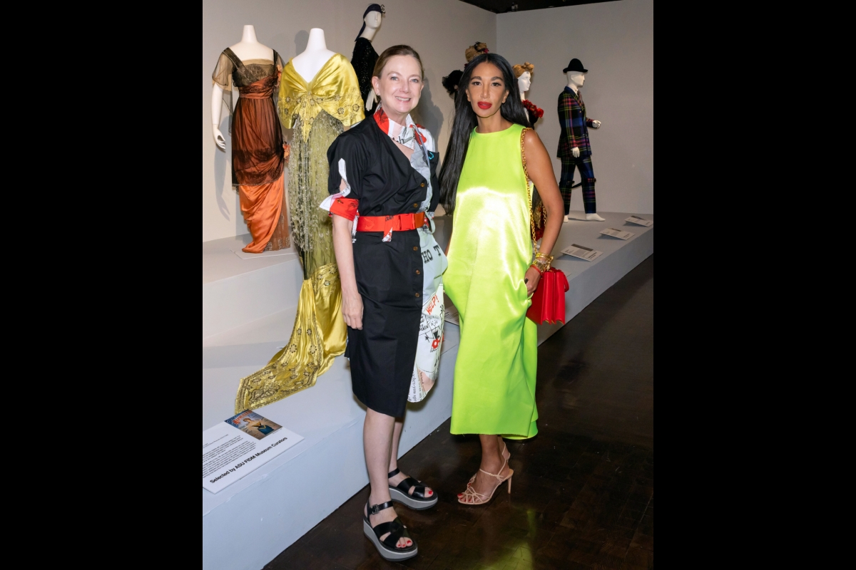 Two women pose next to garments on display in an exhibit.