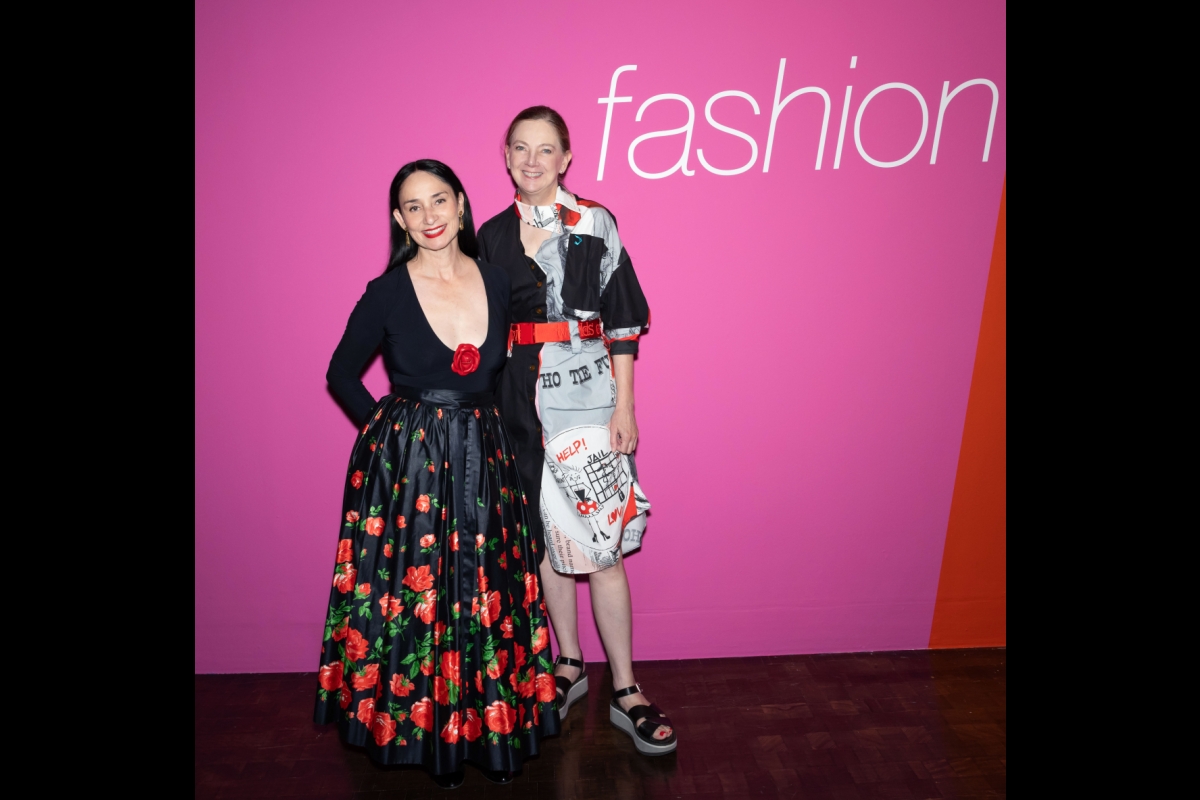 Two women stand in front of a pink wall displaying the word 