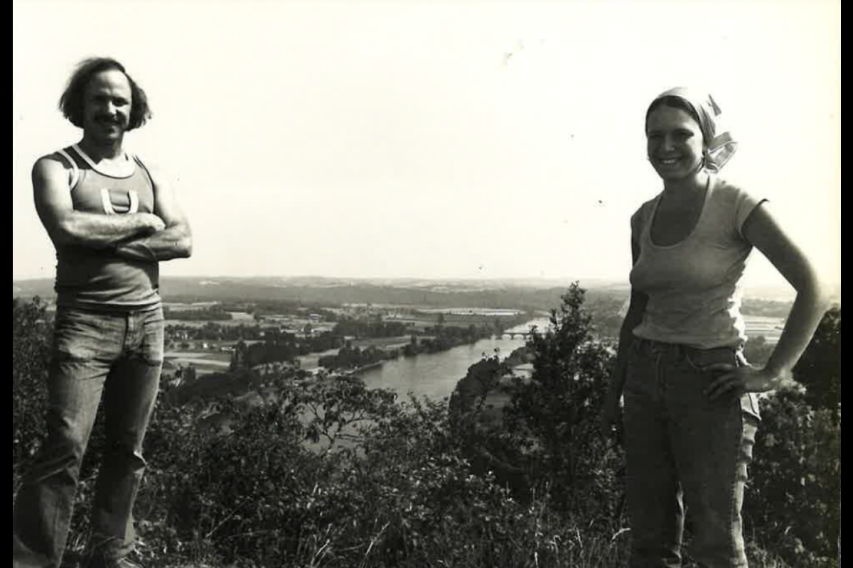 Black-and-white photo of a man and a woman standing in an outdoor setting with a river and cityscape seen in the background.