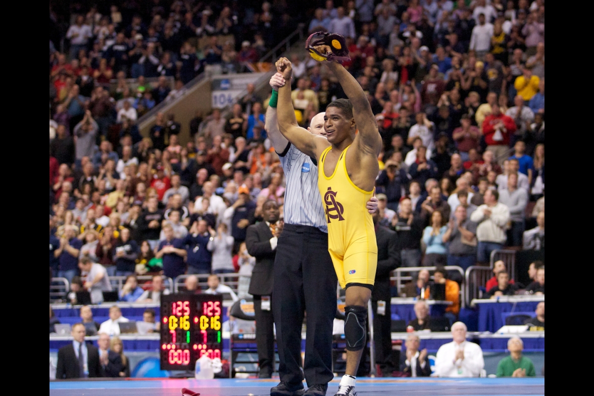 Wrestler in gold uniform with one leg holds hands up victoriously after winning match