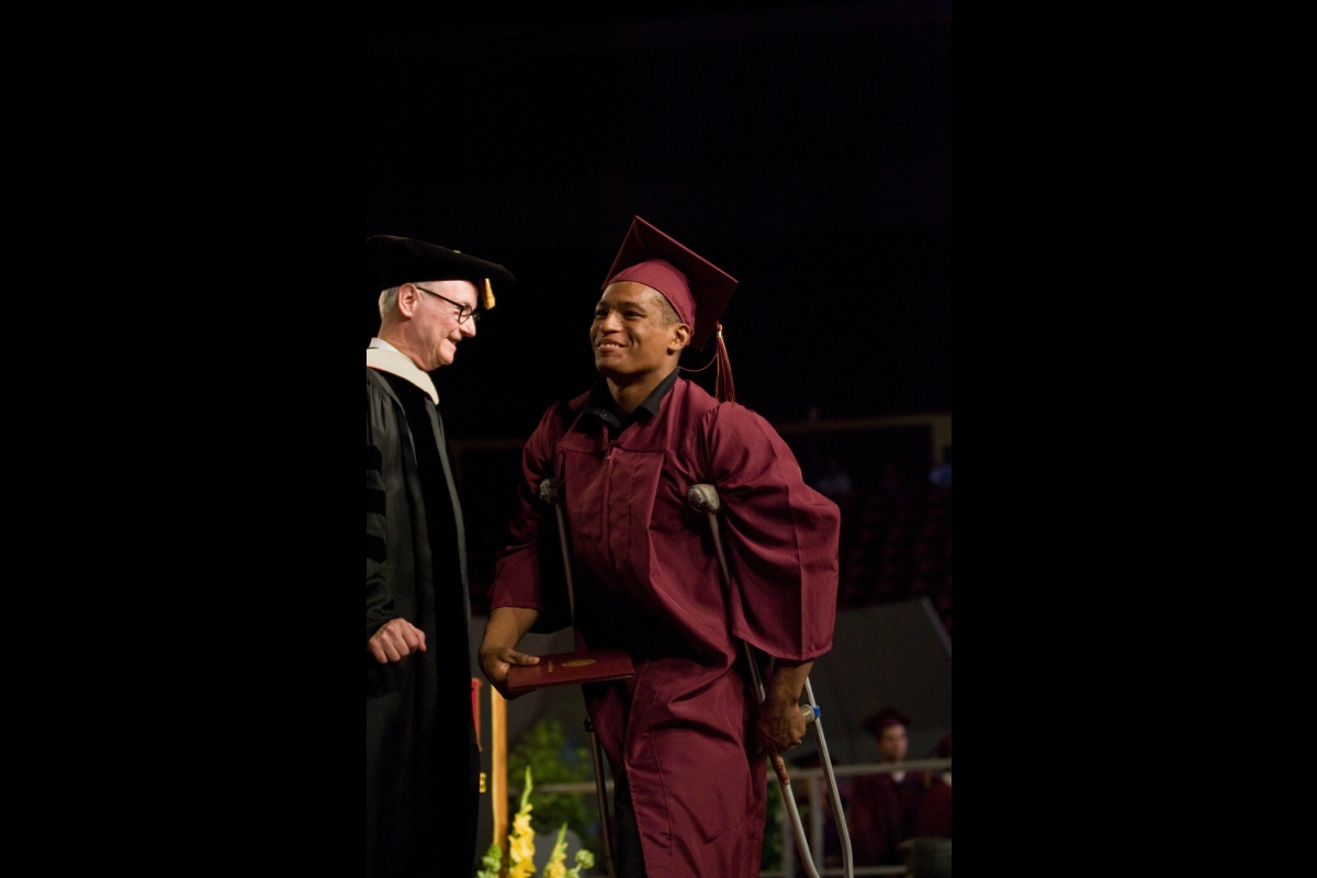 Student in maroon cap and gown crosses stage while using crutches