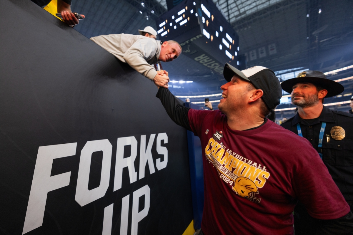 ASU football coach on the sidelines reaches up to clasp hands with ASU basketball coach in stands