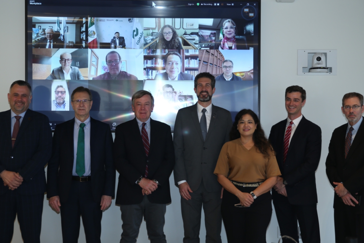 A group photo with seven people standing in front of a screen displaying a Zoom call of several participants.