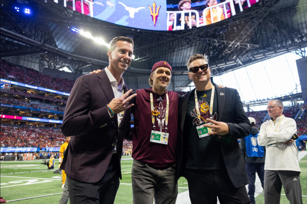 Three men pose for a photo on the sideline of a football stadium