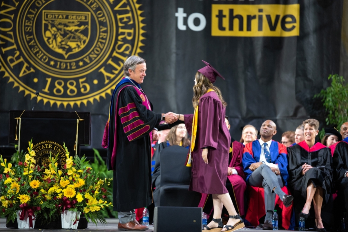 A man in academic regalia shakes the hand of a woman wearing cap and gown as she crosses the stage