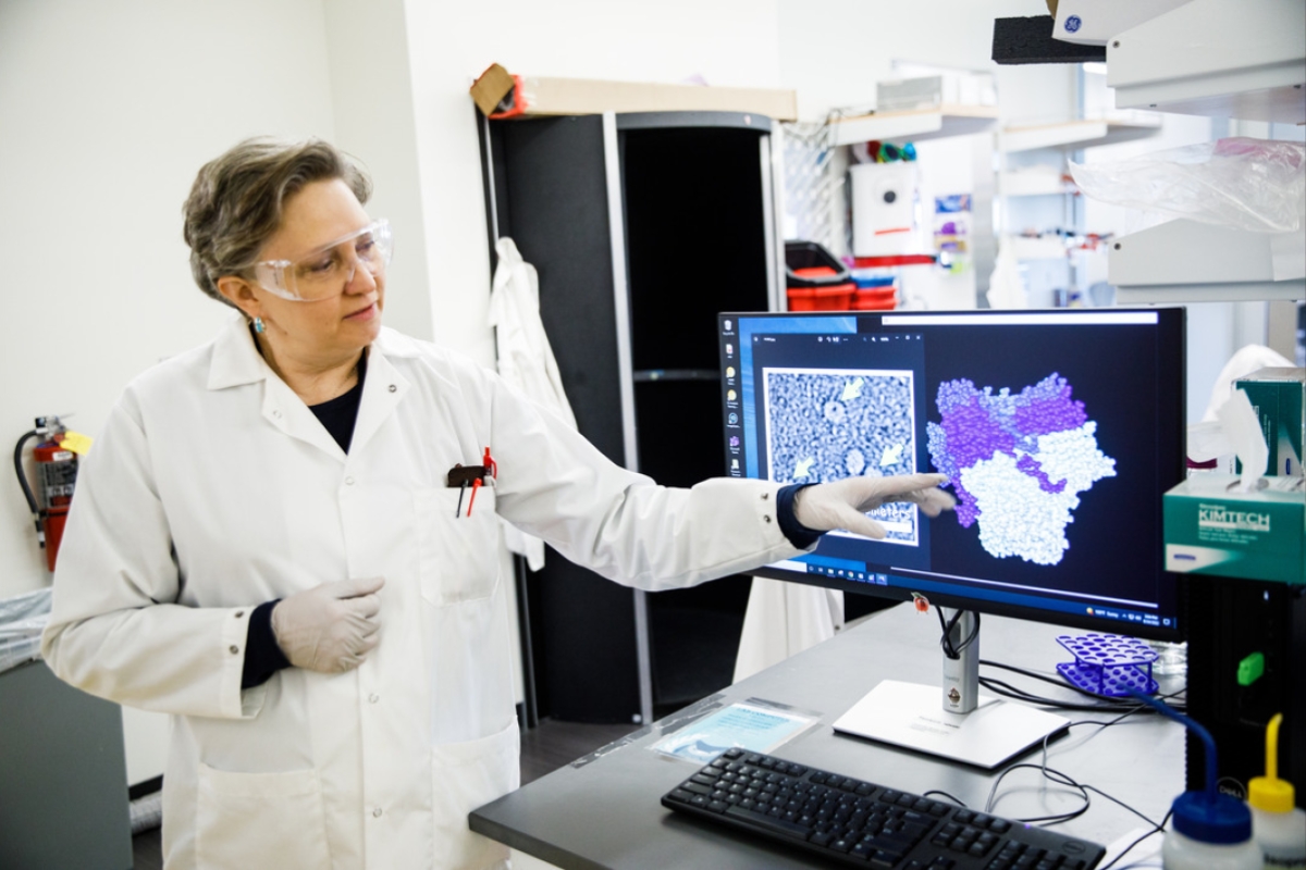 Woman in a white lab coat and goggles points to an image on a computer screen in a lab.
