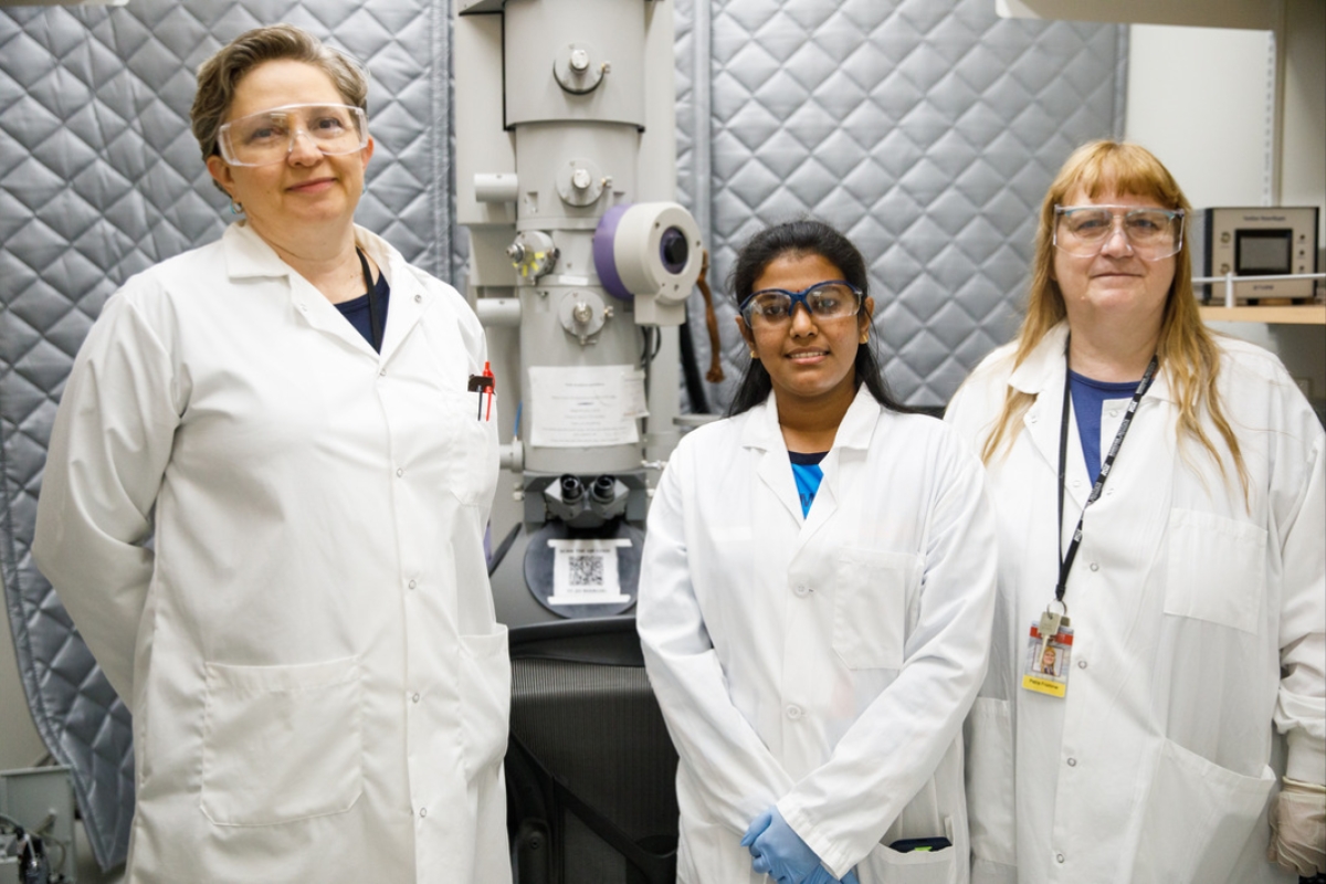 Three women wearing white lab coats and goggles standing in a lab.
