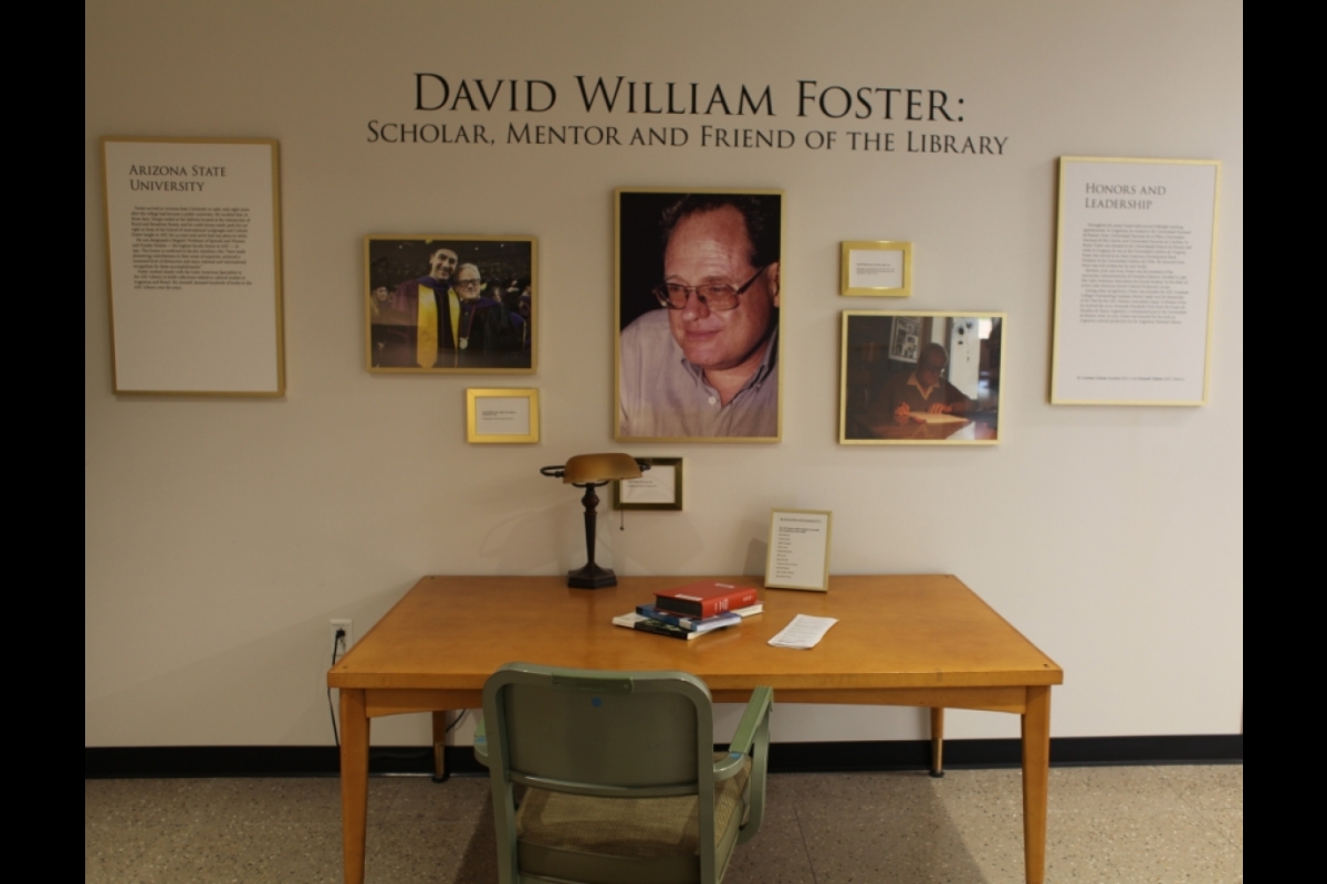 A tan rectangular desk sits in front of a white wall covered in gold framed photos of David William Foster, as well as several signs detailing his life and career. His name is written in large letters on the wall. A lamp and several books sit on the desk.