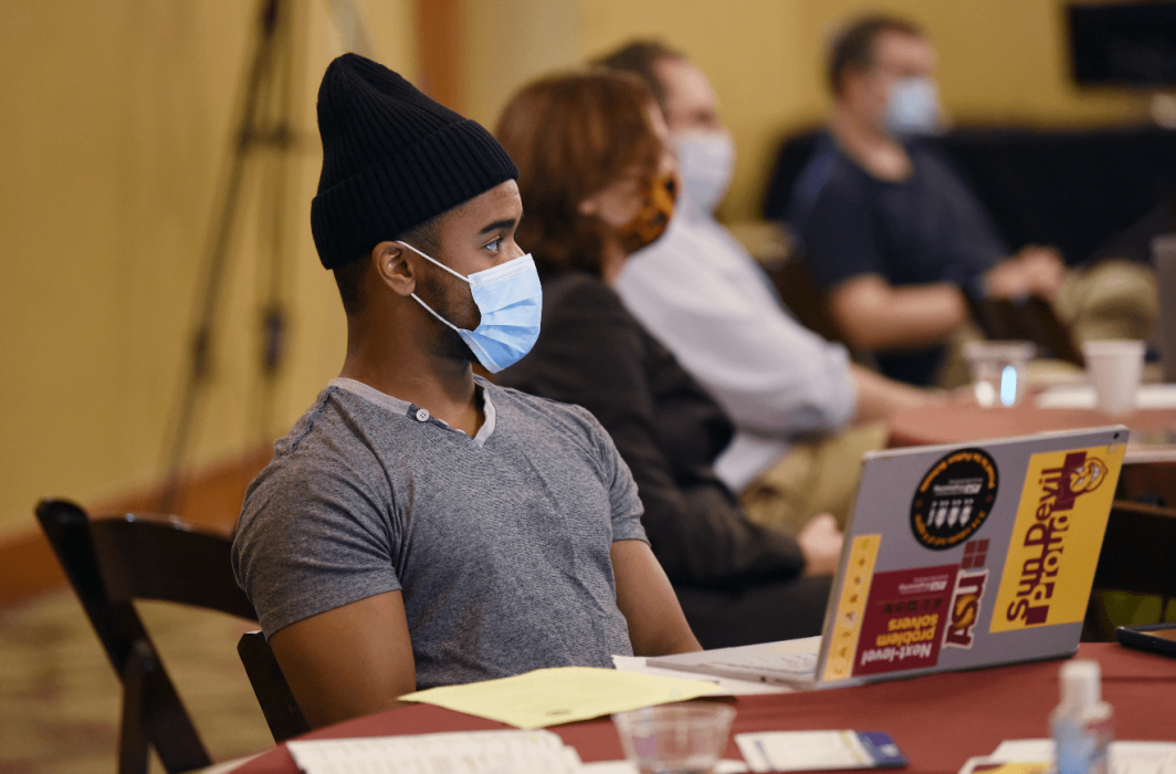 A young Black man listens to a panel discussion. He is wearing a gray V-neck shirt, a black beanie and a blue surgical face mask. He is sitting at a round table with a laptop in front of him covered in maroon and gold ASU stickers. 